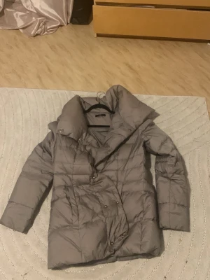 Ljusgrå dunjacka med hög krage - Snygg ljusgrå dunjacka med hög krage och tryckknappar. Jackan har en quiltad design och är fylld med äkta dun för extra värme. Perfekt för kalla vinterdagar och har en modern, något längre passform.