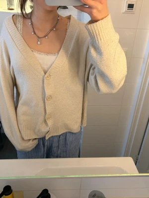 Beige kofta med knappar - Mysig beige kofta med v-ringning och tre knappar framtill. Koftan har långa ärmar och ribbade muddar vid ärmslut och nederkant. Perfekt för lager på lager och enkel att matcha med olika outfits.