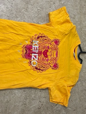 Gul Kenzo t-shirt med tigertryck - Cool gul t-shirt från Kenzo med det ikoniska tigertrycket i rosa och lila på bröstet samt texten 'KENZO PARIS'. T-shirten har rund hals och korta ärmar. Perfekt för dig som vill sticka ut med färg och mönster.