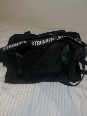 Svart handväska från Stronger - Svart handväska från Stronger med tydlig logotyp på axelremmen i vitt. Väskan har dragkedja, flera bärhandtag och innerfoder med texten 'Escape Ordinary'. Tillverkad i slitstarkt syntetmaterial och har en sportig look.