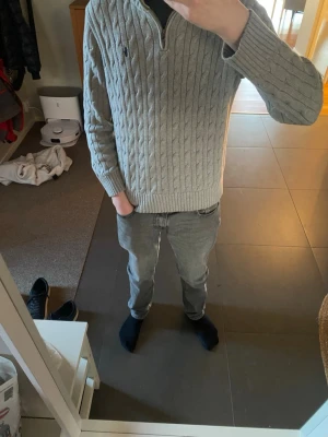 Grå kabelstickad tröja Polo Ralph Lauren - Snygg grå kabelstickad tröja från Polo Ralph Lauren med quarterzip och broderad logga på bröstet. Tröjan har ribbade muddar och hög krage, perfekt för lager på lager. Klassisk och tidlös design som passar till det mesta.