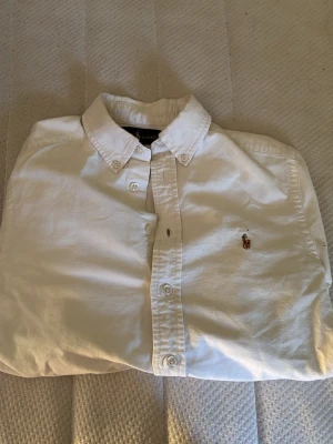Vit skjorta från Ralph Lauren - Klassisk vit skjorta från Ralph Lauren med button-down krage och broderad logga på bröstet. Skjortan har lång ärm, normal passform och stängs med knappar framtill. Perfekt till både jeans och chinos.
