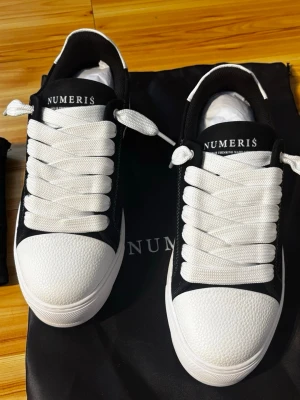 Svarta och vita sneakers från Numeris - Stilrena sneakers från Numeris i svart mocka med vita detaljer och grovvit sula. Skorna har breda vita skosnören, vit tå i texturerat material och en unik sicksack-mönstrad yttersula. Perfekta för dig som gillar en clean och modern look.