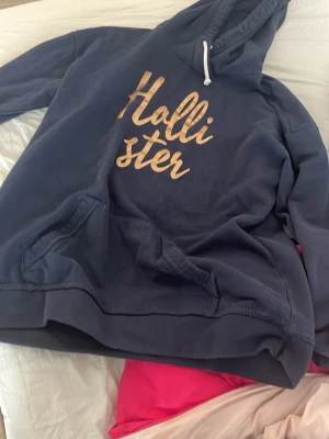 Mörkblå Hollister hoodie - Mysig mörkblå hoodie från Hollister med stor magficka och huva med vit dragsko. Stor logga i guldtext på bröstet ger en snygg och avslappnad vibe. Perfekt för chill dagar eller när du vill ha en casual look.