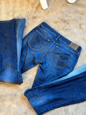 Blå bootcut jeans från Levi's - Säljer ett par klassiska blå bootcut jeans från Replay med fem fickor och snygga slitningar. Jeansen har låg midja, utsvängda ben och detaljerade sömmar på bakfickorna. Perfekta för dig som gillar retrostil och vill ha ett par jeans med skön passform.