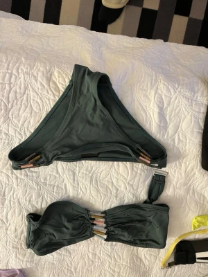 Mörkgrön bikini med detaljer - Snygg mörkgrön bikini med bandeau-topp och matchande trosa. Båda delarna har dekorativa metallfärgade detaljer framtill. Toppen har en axelband och rynkade partier för extra stil. Perfekt för stranden eller poolen!