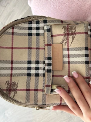 Rutig ryggsäck från Burberry - Säljer en klassisk rutig ryggsäck från Burberry i beige, svart, vitt och rött. Väskan har det ikoniska Burberry-mönstret, detaljer i läder och guldfärgade dragkedjor. Framtill finns en ficka med Burberry-logga och ryggsäcken har ett smidigt handtag upptill.