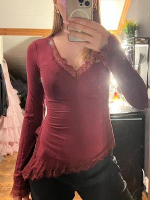 Vinröd långärmad spetsblus - Säljer en vinröd långärmad blus med v-ringning och spetsdetaljer vid halsringning, ärmslut och nederkant. Toppen har en slim passform och är tillverkad i ett mjukt, stretchigt material som sitter snyggt på kroppen. Använd 1 gång. 