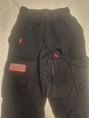 Svarta byxor med cargo-fickor - Svarta byxor från Nike Jordan med stora cargofickor och dragsko i midjan. Byxorna har röda detaljer på snörena och en röd patch på ena benet. Tillverkade i ett lätt, sportigt material och har justerbara benslut.