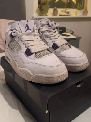 Air Jordan 4 Retro vit/lila - Säljer ett par Air Jordan 4 Retro sneakers i vitt med lila detaljer och meshpaneler. Klassisk siluett med synlig Air-enhet i sulan och Jumpman-logga på plösen. Skorna har vita skosnören och kontrasterande lila inslag vid snörningen.