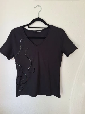 Svart t-shirt med paljettdetaljer från Lindex - Oanvänd svart t-shirt med v-ringning och korta ärmar. På vänster sida finns dekorativa paljettdetaljer i ett slingrande mönster som ger toppen en cool touch. Perfekt att styla med jeans eller kjol för en trendig look.
