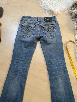 Blå bootcut jeans från Miss Me - Säljer ett par blå bootcut jeans från Miss Me med slitningar och snygga detaljer på bakfickorna. Jeansen har dekorativa sömmar och broderier på fickorna samt en låg midja. Perfekta för dig som gillar en avslappnad men trendig stil. Se bilder för defekter!
