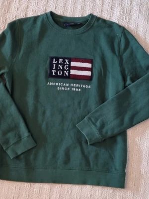 Grön sweatshirt från Lexington storlek 176 - Mörkgrön sweatshirt från Lexington med broderad patch framtill i marinblått, vitt och vinrött. Texten 'AMERICAN HERITAGE SINCE 1993' under patchen. Klassisk rund hals och ribbade muddar vid ärmslut och nederkant. Perfekt för en avslappnad stil. Storlek 176 