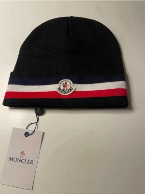 Svart Moncler mössa med ränder - Snygg svart mössa från Moncler med klassiska ränder i blått, vitt och rött samt Moncler-logga framtill. Mössan är ribbstickad och har en uppvikt kant. Perfekt accessoar för att hålla värmen och samtidigt se trendig ut.