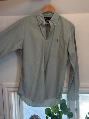 Ljusblå skjorta från Ralph Lauren - Klassisk ljusblå skjorta från Ralph Lauren med button-down krage och broderad logga på bröstet. Skjortan har långa ärmar och vita knappar framtill. Perfekt till jeans eller chinos för en clean och stilren look.