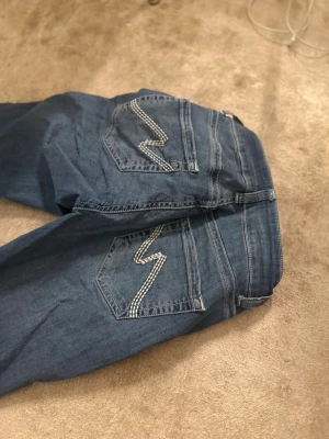 Blå bootcut jeans med mönstrade fickor - Snygga blå bootcut jeans med dekorativa bakfickor som har ett vitt sicksackmönster. Jeansen har klassisk femficksdesign och normalhög midja. Perfekta för dig som gillar en retroinspirerad look och vill ha något som sticker ut lite extra. Ifrån nelly.