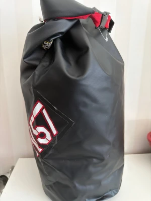 Svart vattentät ryggsäck från Vinga of Sweden - Cool svart ryggsäck från Vinga of Sweden i slitstarkt, vattentätt material. Väskan har en rullstängning med röd detalj och ett stort vitt och rött NS 57-märke på sidan. Perfekt för dig som vill ha en unik och tålig väska till äventyr eller vardag.