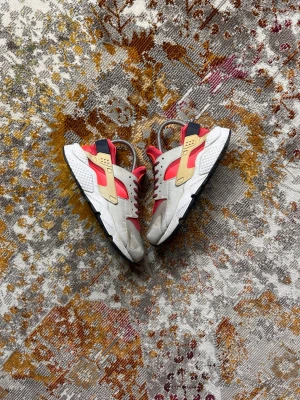 Nike Air Huarache sneakers beige/röd - Nike Air Huarache sneakers med beige och röda detaljer, inslag av svart och gult. Skorna har en chunky sula och en unik design med plastdetaljer på sidorna. Materialmix av läder och mesh, perfekt för dig som gillar statement sneakers.