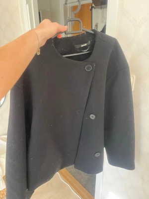 Svart kappa från Bik Bok - Stilren svart kappa från Bik Bok med rak passform och asymmetrisk knäppning framtill. Kappan har rund halsringning och stora svarta knappar. Perfekt för dig som gillar minimalistisk och modern stil.