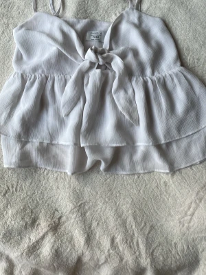 Vit volang topp med knyt framtill - Söt vit linnetopp från NA-KD med tunna axelband, volangdetaljer och knyt framtill. Toppen har en luftig och flowy passform, perfekt för varma dagar. Materialet är lätt och känns skönt mot huden. Nästan helt oanvänd🤗