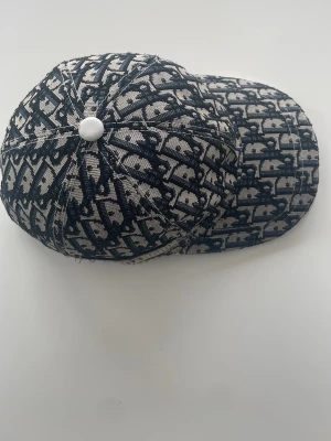 Mönstrad keps från Dior - Säljer en snygg keps från Dior med klassiskt blått och vitt monogrammönster. Kepsen har böjd skärm och är tillverkad i ett tygmaterial med tydlig struktur. Den är inte så gammal har haft denna keps i nog 2 veckor. Pris kan diskuteras !!