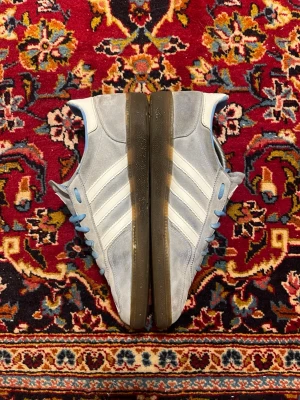 Adidas ljusgrå sneakers med blå detaljer - Säljer ett par ljusgrå Adidas sneakers med klassiska vita stripes på sidorna och blå detaljer vid snörningen och insidan. Skorna har bruna gummisulor.