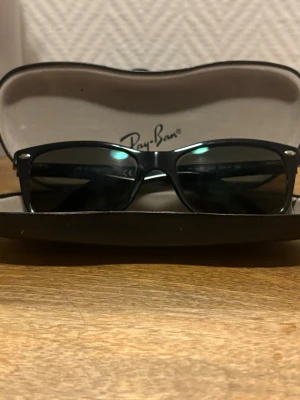 Svarta Ray-Ban Wayfarer solglasögon - Klassiska svarta Wayfarer solglasögon från Ray-Ban med tidlös design och mörka glas. Bågarna är i blank svart plast och har Ray-Ban-loggan på sidan. Perfekta för soliga dagar och ger en cool vibe till din outfit. Dessa solglasögon är rekommenderade för personer som använder glasögon, om det är något du vill byta kan du enkelt byta ut linserna. Dessa köptes för några år sedan för 3000 kr.