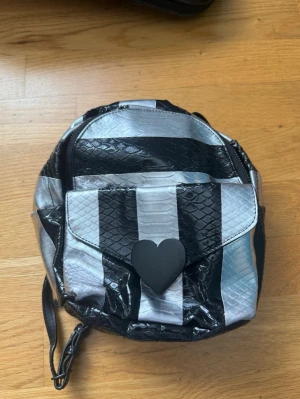 Randig ryggsäck med hjärtdetalj - Säljer en unik ryggsäck i svart och silver med ormmönstrad yta och breda ränder. Framtill finns en ficka med lock och en svart hjärtformad knapp. Axelbanden är justerbara och delvis i kedja. Perfekt för dig som gillar edgy och annorlunda accessoarer.