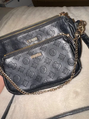 Svart handväska med kedja ”från guess märket” - Säljer en svart handväska från Guess med präglat logomönster och guldfärgade detaljer. Väskan har två fack med dragkedja och en snygg kedjerem. Perfekt för dig som gillar stilrena accessoarer med en touch av lyx.