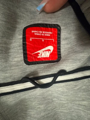 Grå zip hoodie från Nike - Snygg grå zip hoodie från Nike med svarta detaljer och dragkedjor på fickorna. Klassisk passform med huva och ribbade muddar. Perfekt för en sportig och avslappnad stil. Tillverkad i mjukt material för extra komfort. Hål under armhålorna säljer billigare…