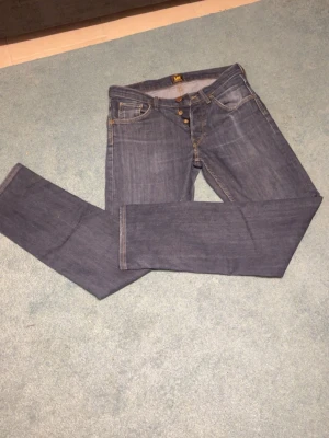 Blå raka jeans från Lee - Säljer ett par klassiska blå jeans från Lee med raka ben och knappgylf. Jeansen har fem fickor och kontrastsömmar i beige. Tillverkade i slitstark denim som passar perfekt till en avslappnad stil.