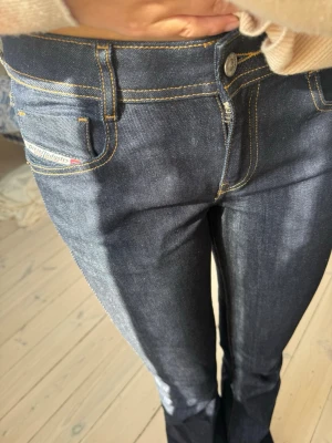 Mörkblå bootcut jeans från Diesel - Säljer ett par mörkblå bootcut jeans från Diesel med klassiska gula sömmar och femficksmodell. Jeansen har låg midja och en liten Diesel-logga på fickan fram. Materialet är kraftig denim som sitter snyggt och formar benen.