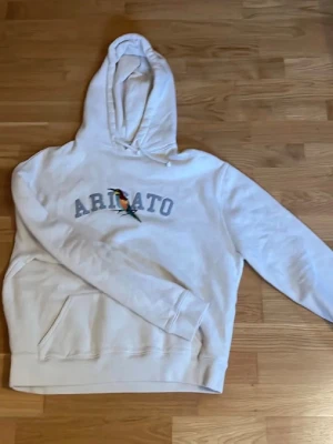 Vit hoodie från Axel Arigato - Säljer en beige hoodie från Axel Arigato med tryckt logga och en färgglad fågel på bröstet. Hoodien har stor huva och en klassisk magficka framtill. Tillverkad i mjukt material, perfekt för en chill och stilren look.