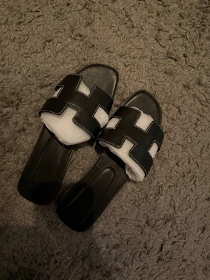 Svarta slip-in sandaler med vit söm - Stilrena svarta slip-in sandaler med breda band i läderlook och kontrasterande vita sömmar. Klassisk design med öppen tå och fyrkantig form på sulan. Perfekta för en trendig och avslappnad stil.