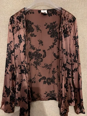 Rosa blommig omlottblus i Silke NoaNoa - Snygg omlottblus i glansigt rosa satin med svarta blommönster. Blusen har långa ärmar och knytband framtill för en cool och trendig look. Perfekt att styla med jeans eller kjol för en chic outfit.