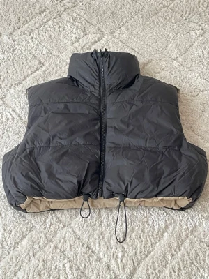 Svart kort dunväst med hög krage - Snygg svart dunväst i boxig, croppad modell med hög krage och dragkedja framtill. Västen har beige detaljer längst ner och justerbar dragsko i nederkant. Perfekt för lager på lager och streetwear-vibe.