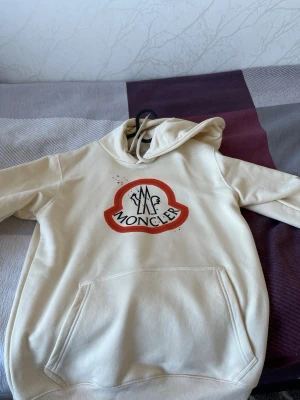 Beige hoodie från Moncler - Snygg beige hoodie från Moncler med stor logga framtill i rött och svart. Klassisk känguruficka och huva med dragsko. Tillverkad i mjukt material som känns skönt mot huden. Perfekt för dig som vill ha en stilren och trendig look. Riktig från moncler och pris kan diskuteras 