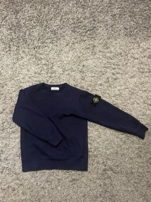 Mörkblå sweatshirt från Stone Island - Säljer en mörkblå sweatshirt från Stone Island med klassisk rund hals och ribbade muddar som är köpt här på Plick! Tröjan har den ikoniska Stone Island-loggan på vänster ärm. Perfekt för en avslappnad och stilren look. Tveka inte på att höra av dig vid minsta fundering.