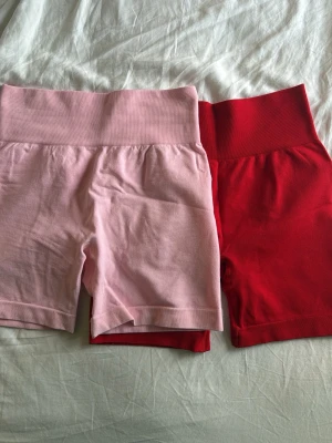 Gymshorts - Säljer två par gymshorts med hög midja, ett par i ljusrosa och ett par i rött. Båda är ribbade i midjan och tillverkade i mjuk bomull, perfekta för en avslappnad och sportig stil.