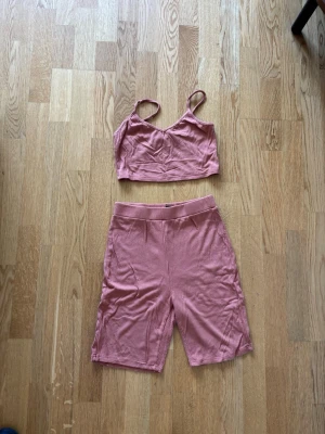 Rosa ribbat set med topp och shorts - Säljer ett rosa ribbat set med en croppad topp med smala axelband och matchande shorts med resår i midjan. Setet är mjukt och stretchigt, perfekt för att chilla hemma eller som underkläder. Inget synligt märke.
