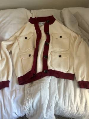 Krämfärgad kofta med vinröda detaljer - Snygg kofta från Gasta Studio i krämvitt bomullstyg med vinröda ribbade muddar och knappslå. Fyra stora fickor framtill med knappar ger en cool utility-vibe. Perfekt till jeans eller kjol för en trendig look.