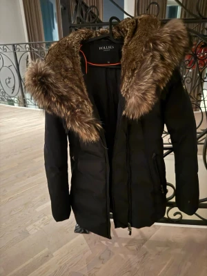 Svart hollies jacka - Stilren svart kappa från Hollies med stor, fluffig päls runt huvan. Lång modell med dragkedja framtill och fickor på sidorna. Perfekt för kalla dagar när du vill vara både varm och snygg. Klassisk design som passar till det mesta.