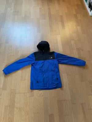 Blå och svart vindjacka The North Face - Snygg blå vindjacka från The North Face med svarta detaljer över axlar och huva. Jackan har dragkedja framtill, två sidofickor och justerbar huva. Perfekt för blåsiga dagar och enkel att matcha med olika outfits. Tillverkad i lätt och tåligt material.