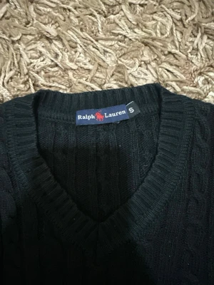 Svart kabelstickad tröja Ralph Lauren - Kabelstickad svart tröja från Ralph Lauren med V-ringning och ribbade muddar. Klassisk design med diskret logga på insidan av kragen. Perfekt till lager på lager och enkel att matcha med olika outfits.