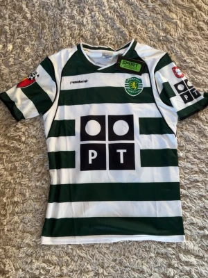 Sporting Lissabon C. Ronaldo matchtröja Reebok - Grönvit randig matchtröja från Sporting Lissabon med tryckta loggor och sponsortryck. Tröjan har korta ärmar, klubbmärke på bröstet och nummer 28 samt C. Ronaldo på ryggen. Tillverkad i lätt och ventilerande material, perfekt för fotboll.