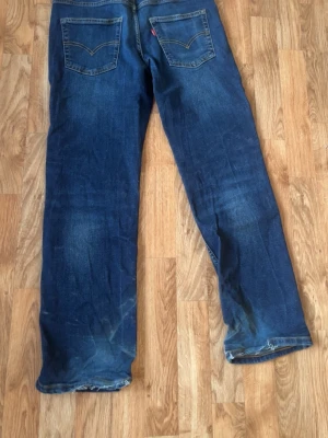 Blå raka jeans från Levi's - Säljer ett par klassiska blå jeans från Levi's med raka ben och femficksmodell. Jeansen har snygga slitningar och kontrastsömmar, samt den ikoniska röda Levi's-lappen på bakfickan. Perfekta för en avslappnad och tidlös stil.