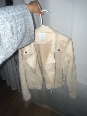 Beige skinnjacka med dragkedjor - Snygg beige skinnjacka från Lindex  med klassisk bikerkrage, silverfärgade dragkedjor och tryckknappar. Jackan har en figurnära passform och är perfekt för dig som vill ha en stilren och trendig look. Har ba använt den en gång!!
