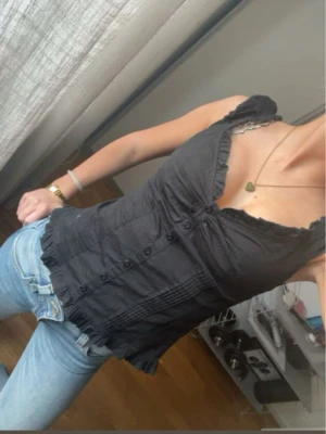 Svart blus med volang och knappar - Supersöt svart blus med volangkant vid ärmar och nederkant. Toppen har v-ringning, dekorativa knappar framtill och smickrande sömmar som ger en feminin siluett. Perfekt att styla med jeans eller kjol för en trendig look.