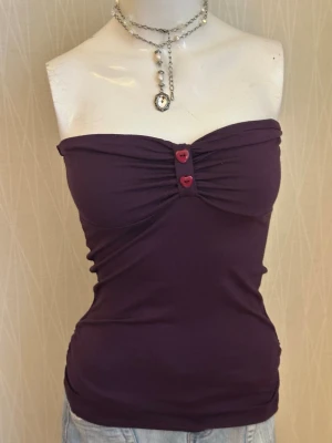 Mörklila tubtopp med knappar - Snygg mörklila tubtopp med två dekorativa röda knappar framtill. Toppen är bandeau-modell och har en tight passform som framhäver figuren. Perfekt att matcha med jeans eller kjol för en trendig look.