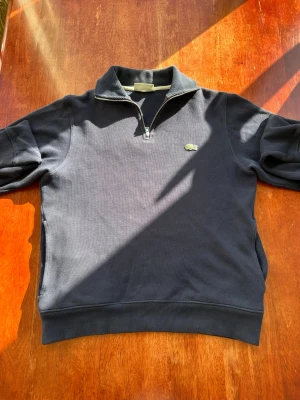 Mörkblå halfzip tröja från Lacoste - Snygg mörkblå tröja från Lacoste med halv dragkedja och klassisk krokodillogga på bröstet. Tröjan har ribbade muddar och är tillverkad i mjukt bomullsmaterial. Perfekt för en clean och stilren look.